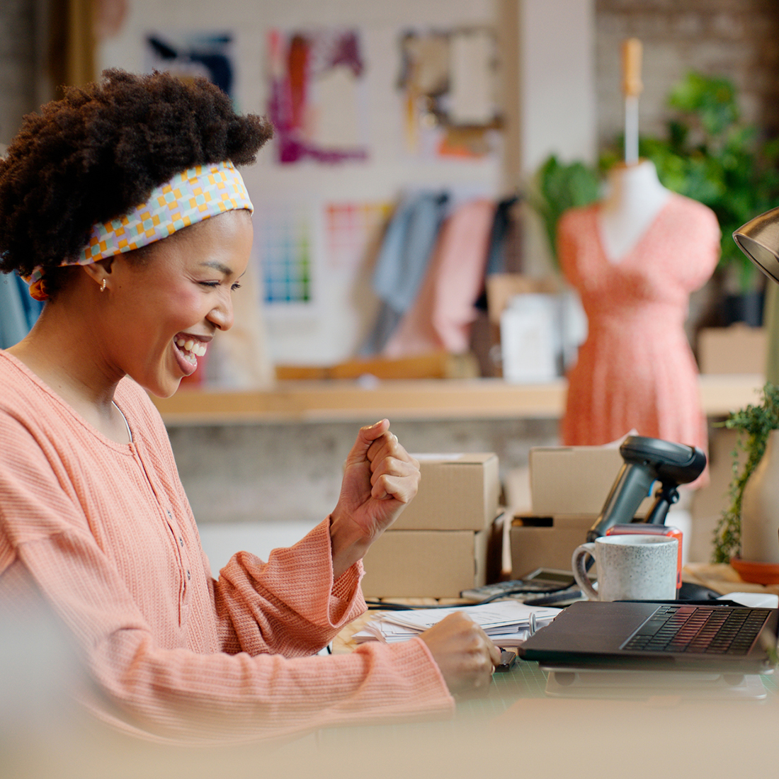 Femme souriante dans un atelier de couture avec ordinateur portable et boîtes d'expédition sur la table.