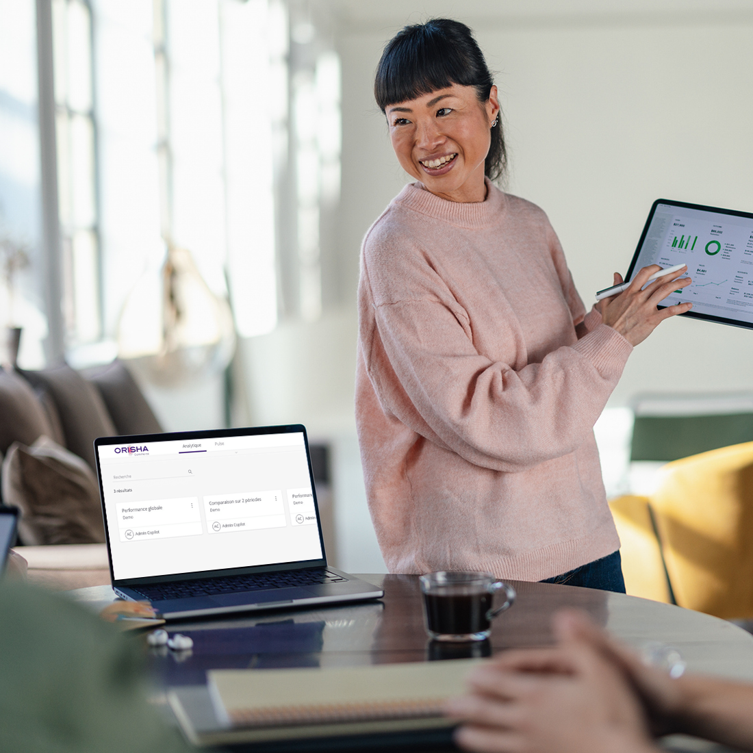 Femme souriante présentant des données statistiques sur une tablette devant un ordinateur portable.