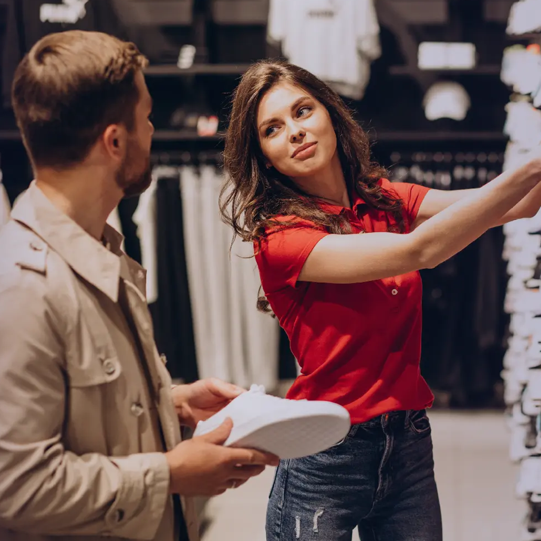 Une vendeuse en rouge montre un vêtement à un client tenant une chaussure blanche en magasin.