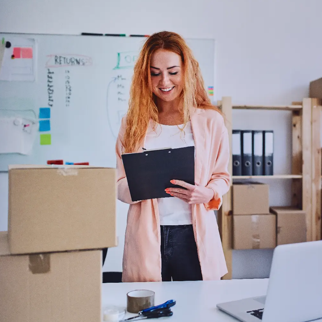 Femme souriante examinant un tableau avec des boîtes en carton et un ordinateur portable sur le bureau.