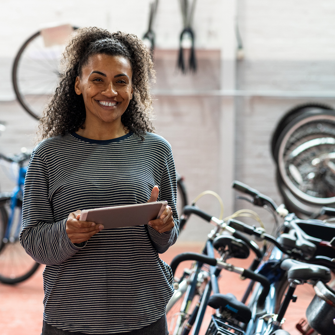 Femme souriante avec tablette dans un atelier de réparation de vélos avec plusieurs bicyclettes autour.