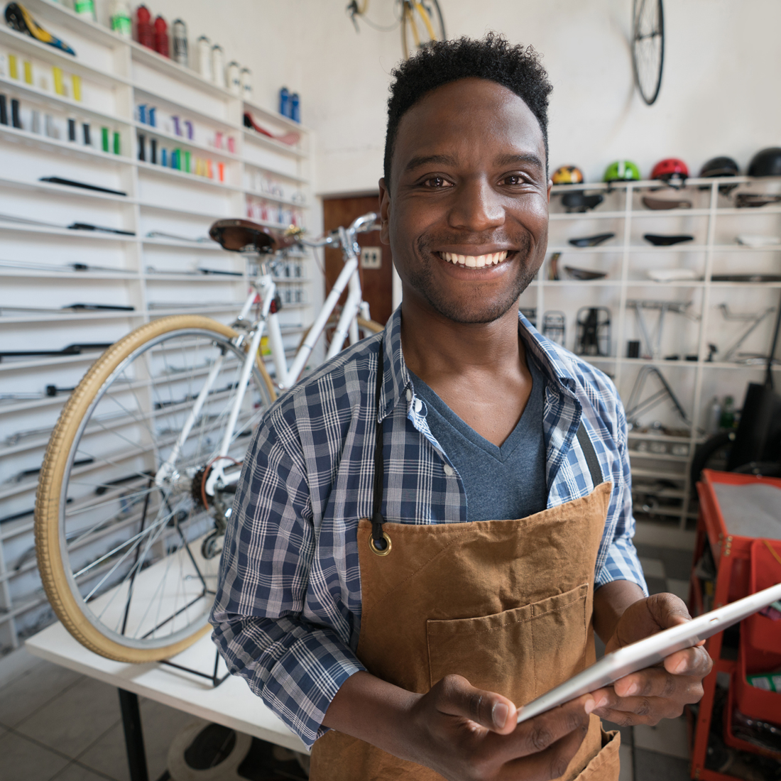 Un homme souriant portant un tablier marron dans un atelier de réparation de vélos avec une tablette.