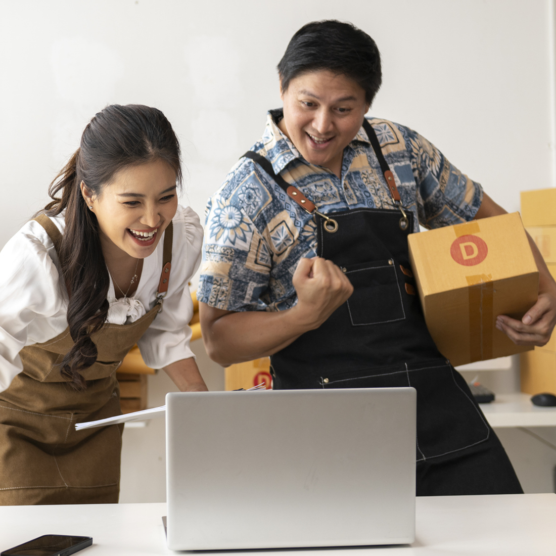 Deux collègues souriants regardant un ordinateur portable, un tenant une boîte en carton et l'autre des papiers.