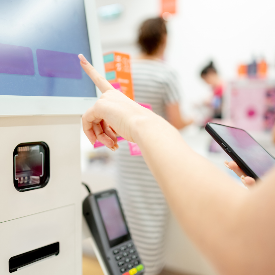 Main visible hand taps a touchscreen on a self-checkout kiosk holding a smartphone in the other hand.