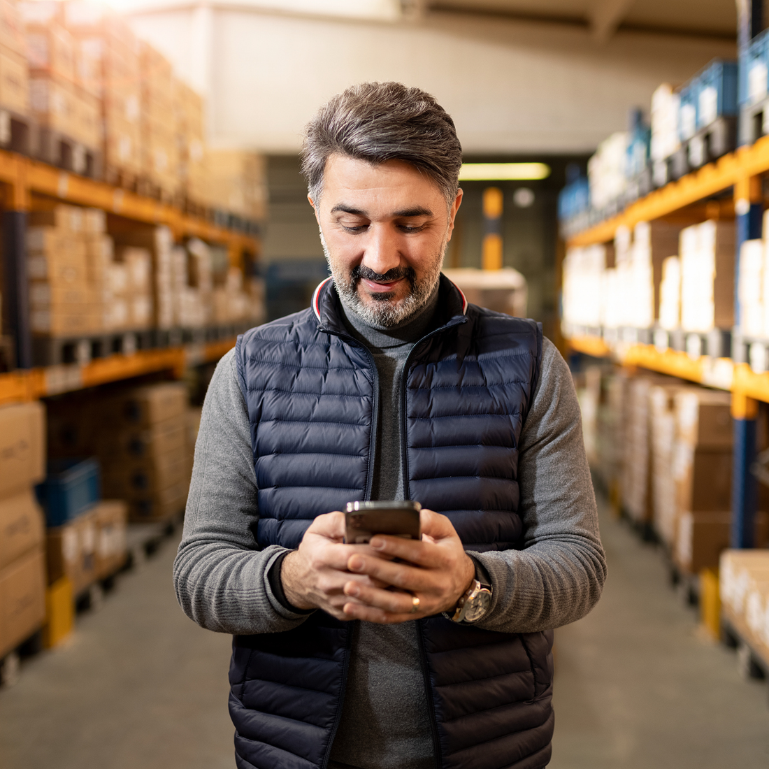 Un homme souriant consulte son téléphone portable dans un entrepôt rempli de boîtes.