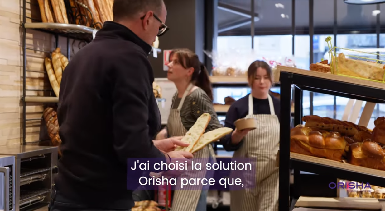Un homme tenant du pain dans une boulangerie avec deux employées en arrière-plan.
