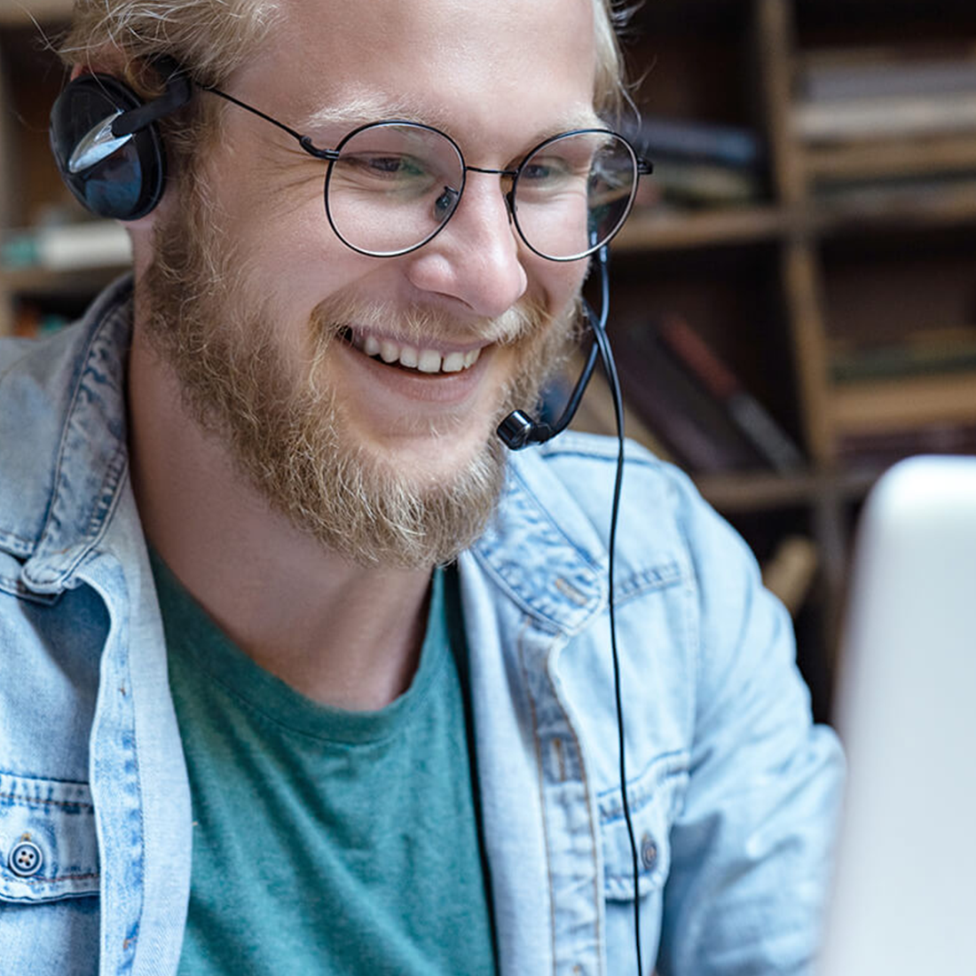 Jeune homme souriant avec casque micro et lunettes, devant un ordinateur portable.