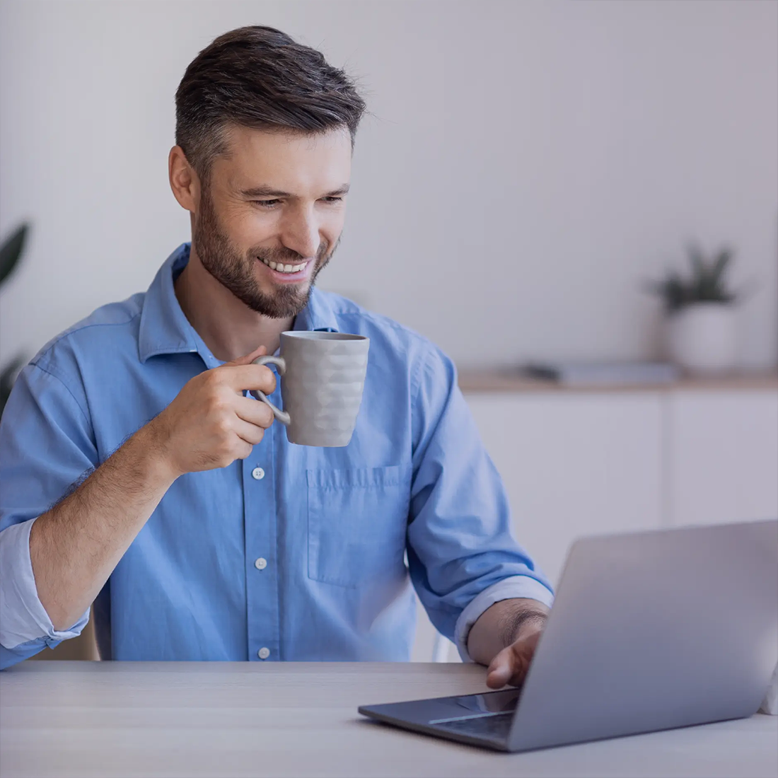 Homme souriant en chemise bleue tenant une tasse et regardant un ordinateur portable.