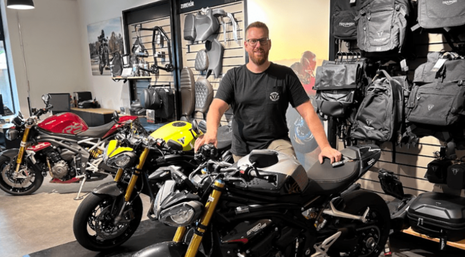 Un homme se tient dans un magasin de motos avec trois motos et des sacs à dos Triumph accrochés au mur.