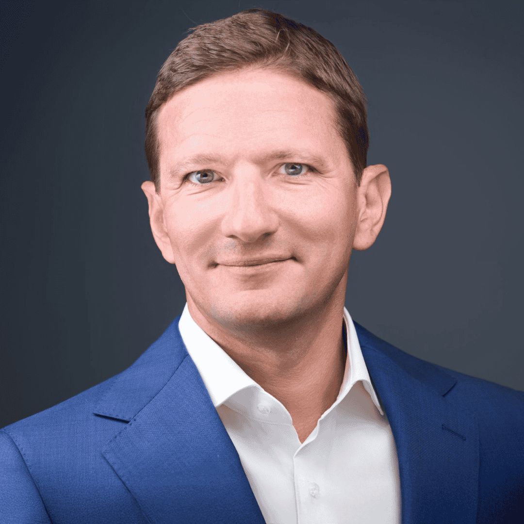 Homme souriant en costume bleu et chemise blanche devant un fond gris foncé.