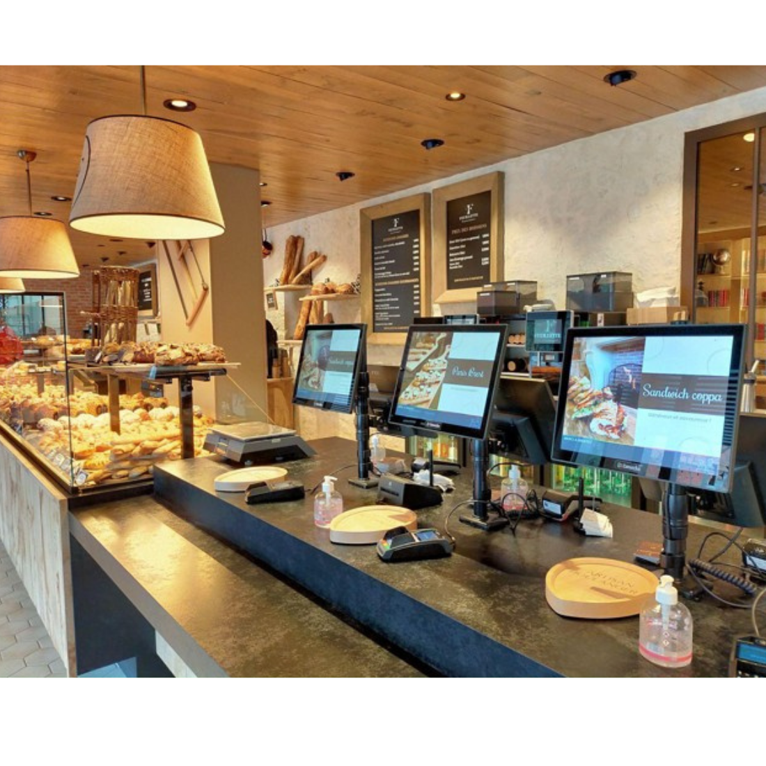 Comptoir d’une boulangerie avec caisses enregistreuses, écrans, pains, viennoiseries et lampes suspendues.