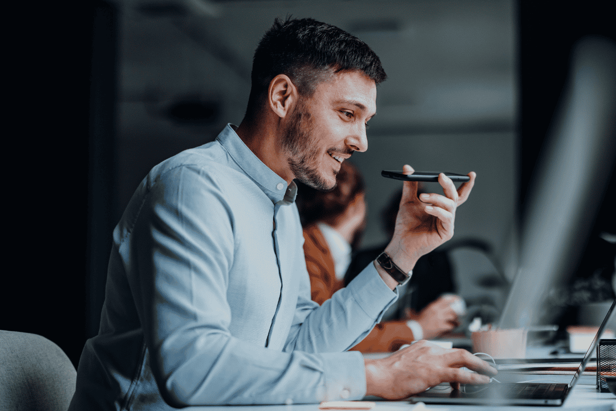 Homme parlant dans un smartphone en mode haut-parleur tout en utilisant un ordinateur portable au bureau.