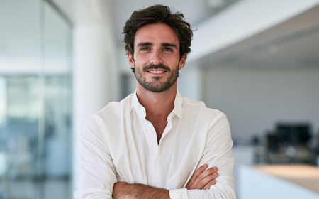 Homme souriant avec une chemise blanche, bras croisés dans un bureau moderne et lumineux.