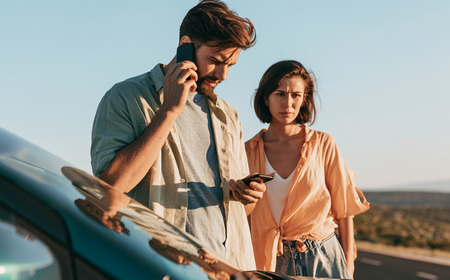 Un homme parle au téléphone portable tout en regardant un autre téléphone, une femme debout à côté de lui semble préoccupée, tous deux près d'une voiture garée sur le bord d'une route en plein air avec un ciel clair.