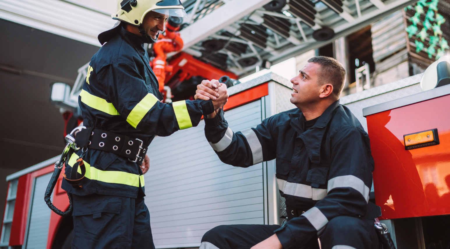 Deux pompiers en tenue de protection noire à bandes réfléchissantes, l'un debout en casque serre la main de l'autre assis près d'un camion de pompiers rouge lors d'une pause ou d'une communication.